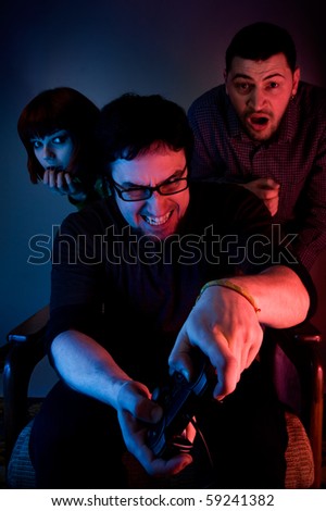 Young emotional man playing video games, two scared persons are hiding behind him