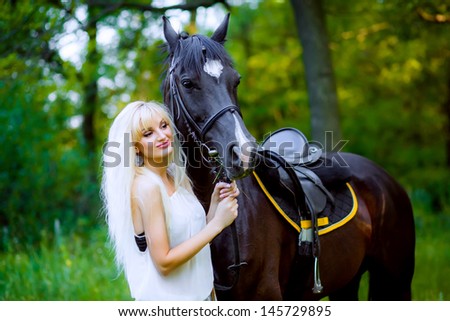 Beautiful young blonde woman sitting on a horse in white dress, in green garden.
