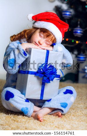 Happy red-haired girl in a Christmas cap sits near a Christmas tree at home and went to sleep hugging a gift