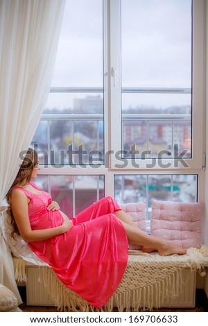 beautiful pregnant woman sitting on the window in the room and looks into the distance