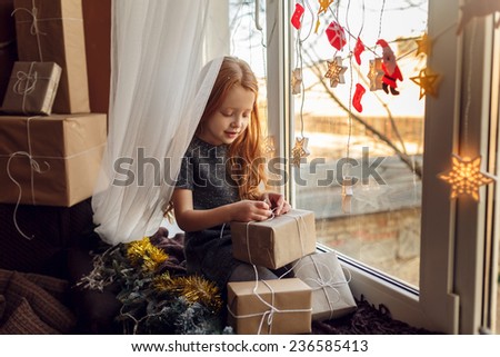 Adorable little girl with red hair in a room near the window with gifts. Christmas holidays. In anticipation of the holiday