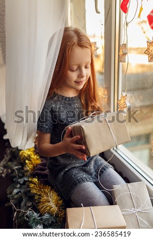 little girl with red hair in a room near the window with gifts. Christmas holidays, children\'s emotions. In anticipation of the holiday