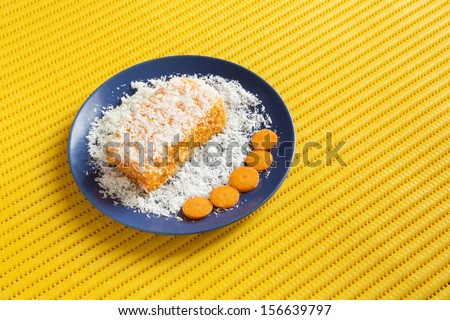 Piece of carrot and coconut cake with some layers of cookies, covered with grated coconut in a blue plate with some carrot slices on a yellow plastic tablecloth full of small holes