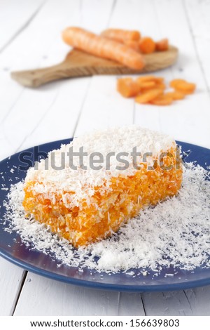 Piece of carrot and coconut cake with some layers of cookies, covered with grated coconut in a blue plate on a white weathered table with some carrots as background