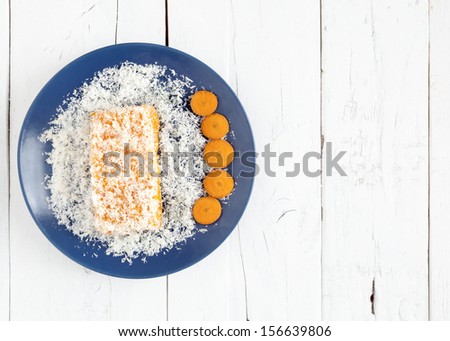 Piece of carrot and coconut cake cover with grated coconut in a blue plate with some carrot slices on a white weathered table with copy space