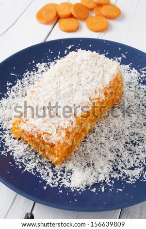 Piece of carrot and coconut cake with some layers of cookies, covered with grated coconut in a blue plate on a white weathered table with some carrot slices as background