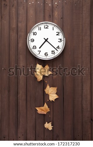Wall clock hanging on the top in a brown plank with some dry leaves pretending a pendulum below it
