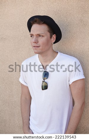 Young adult man looking away. He stands on a textured yellow wall and he wears a hat, glasses and a white t-shirt.