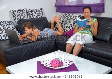 Little girl using computer in the living room while her mother is reading a book