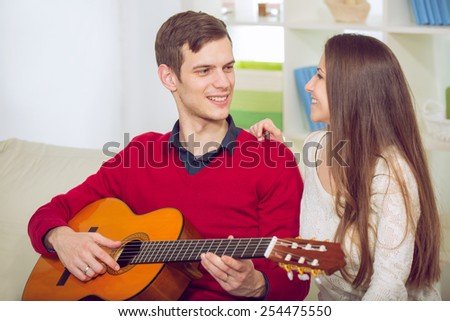 A young and attractive couple sitting on a couch with a guitar. The male is playing the guitar. Selective focus