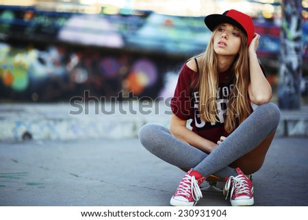 Portrait of beautiful teen girl sitting on skateboard over wall with abstract graffiti art. Urban outdoors, teenager\'s lifestyle