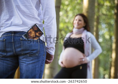 A man stands facing his pregnant woman. Ultrasound picture of baby in his pocket. She is standing in the background and out of focus.Focus on ultrasound image.