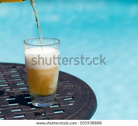 Plain pint glass of beer being poured sitting on table by blue swimming pool