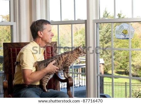 Senior man with bengal cat watching chickadee bird on birdfeeder from chair