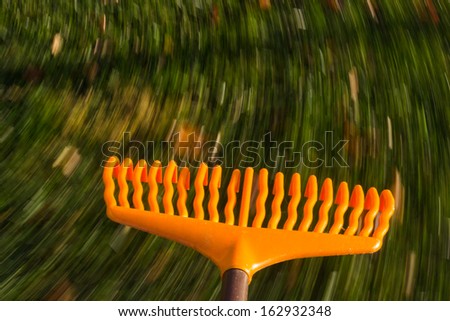 Motion blur created when orange lawn rake is moved through the leaves on green lawn with a focus on the tool as the grass and fallen autumn leaves blur with movement to suggest work