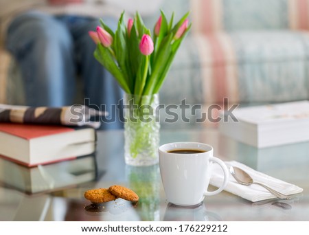 Modern White Porcelain Cup Of Black Coffee On Glass Table With Spoon And Ginger Biscuits. Book Newspaper And Vase Of Tulips In The Background Out Of Focus