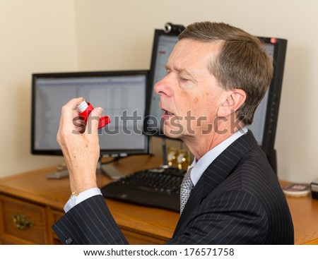 Senior caucasian man  in suit at desk with computer screens with asthma inhaler to handle problems with breathing