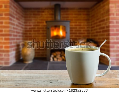 Coffee cup on wooden table in front of roaring fire inside wood burning stove in brick fireplace