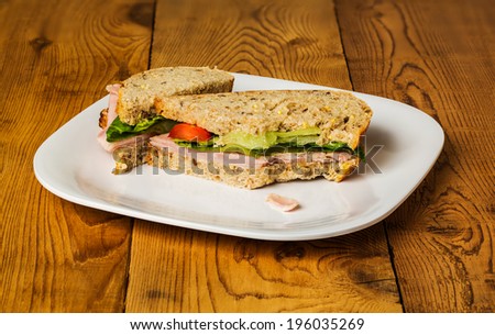 English ham lettuce and tomato sandwich with a bite out of one corner. Bread is artisan multigrain wheat. Second sandwich is behind the first and both are on white picnic plate on rough wooden table