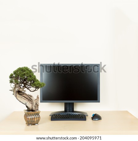 Small old bonsai tree in golden pot on plain wooden desk with computer monitor and keyboard to suggest calm and meditation at work