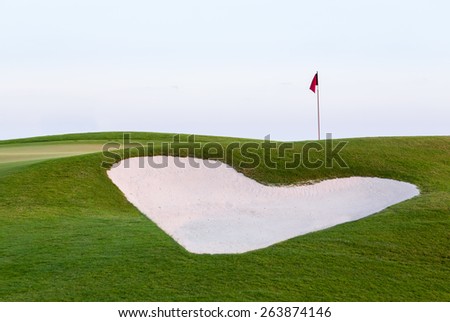 Heart shaped sand bunker in front of red flag of golf hole on beautiful course at sunset illustrating love for game of golf