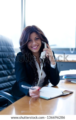 young business woman sitting at the desk talking at the phone on office background