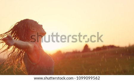 CLOSE UP, LENS FLARE, COPY SPACE: Smiling young woman spins in the rain with her arms outstretched. Blonde haired Caucasian girl playfully dances at stunning sunset. Female dancer improvising in rain