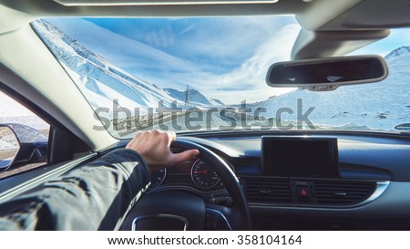 view from luxury car inside with part of interior gps screen with driver male hand on  the steering wheel during bright snowy sunny day on straight ice road with snowy mountains in background