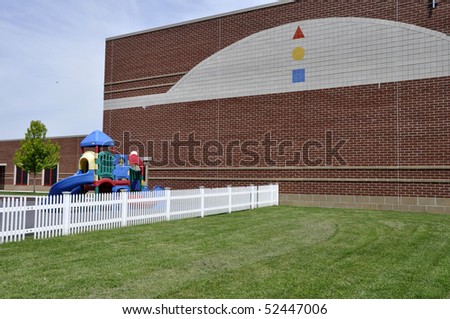 Playground and a white fence by a modern red brick school building.  There is a bright green lawn by the fence.