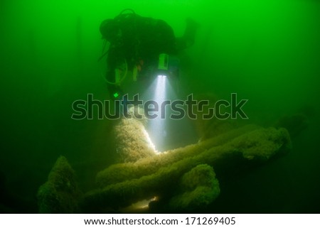 Using a light, a diver explores one of the many shipwrecks found off of Cape Cod, Massachusetts. The green, plankton-filled water here is extremely cold and thus the diver is outfitted in a drysuit.
