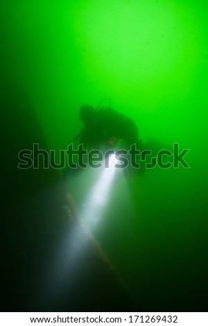 Using a light, a diver explores one of the many shipwrecks found off of Cape Cod, Massachusetts. The green, plankton-filled water here is extremely cold and thus the diver is outfitted in a drysuit.