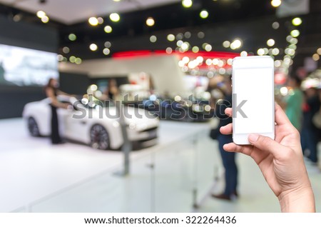 Female hand holding mobile smart phone on the Abstract blurred photo of motor show, car show room