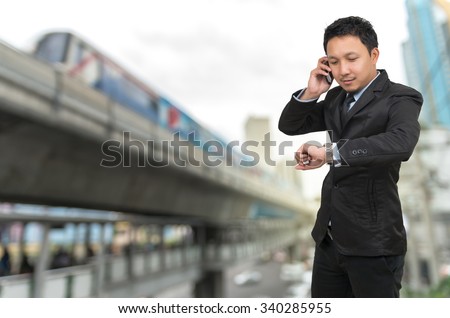 Young businessman talking on mobile phone and looking at watch on abstract Blurred photo of sky train with traffic and cityscape, business rush hour concept