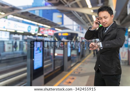 Young businessman talking on mobile phone and looking at watch on Abstract blurred photo of sky train station with people background, business rush hour concept
