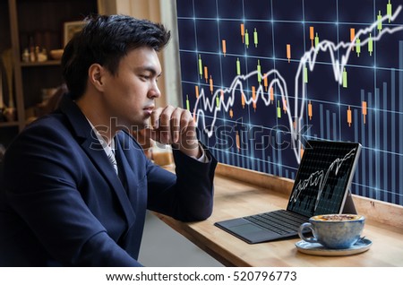 Asian Businessman sitting and using computer laptop showing trading graph beside the windows glass over the stock exchange trading graph screen background, Business financial and forex concept