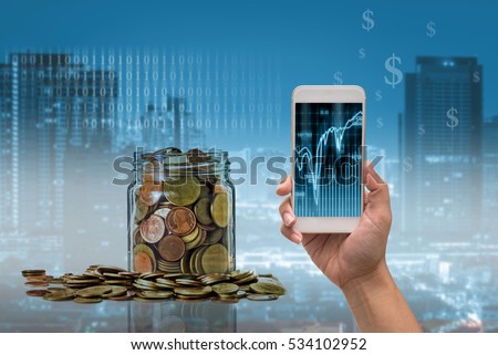 Female hand holding mobile smart phone touch screen showing the stock market chart over the Gold coins in clear bottle on Trading graph with cityscape, Business financial and technology concept