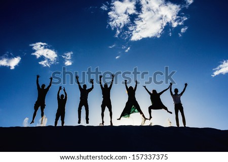 Black silhouettes of seven enthusiast people jumping on sand. Blue sky behind.