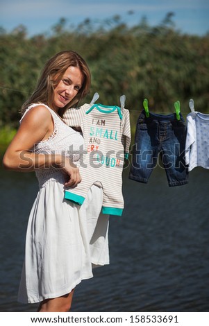 Young pregnant woman is holding baby clothes from laundry line next to a river