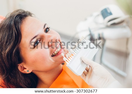 Young smiling woman lying on dentist\'s chair and determining teeth\'s color  using shade guide