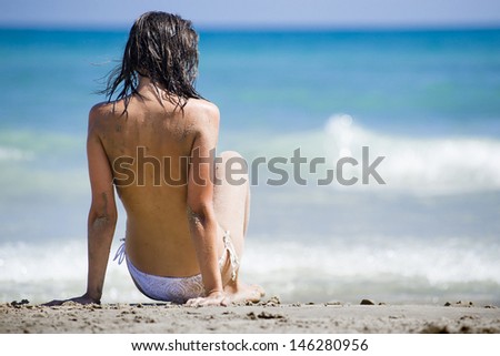 Topless Woman sitting with her back facing the camera, in the sand wearing a white bikini with the sea in the back ground