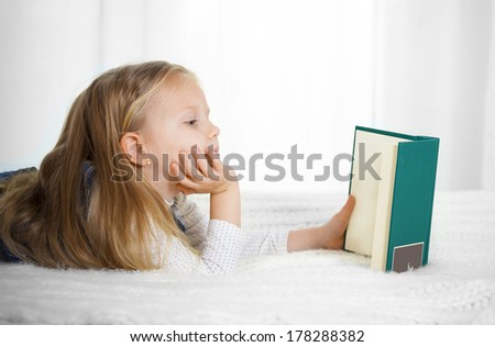 happy cute blonde haired school girl wearing a school uniform reading a book lying on the bed