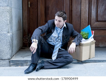 desperate business man in stress fired from job sitting on edgy street corner with office belongings in cardboard box and asking for help