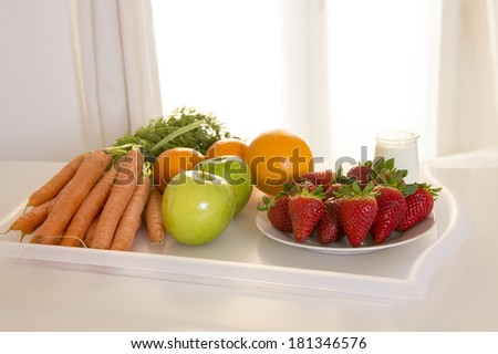 clear tray of fresh fruit and vegetable, carrots, oranges, apples and strawberries backlight in front of a white window