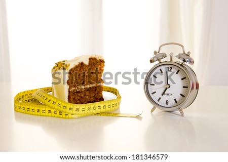 carrot cake wrapped in a measuring tape with a alarm clock, a concept to remind people when to start and not eat junk sugar foods backlit with a white window in the background