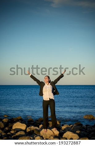 happy attractive business woman at the beach wearing suit away from office standing on some rocks with her hands in the air watching and enjoying the sunset.