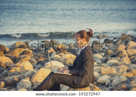 attractive business woman working on computer sitting on some rocks at the beach looking up to sky thinking about freedom