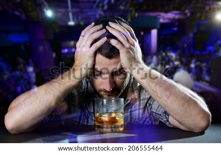 young alcoholic drunk man thoughtful about alcohol addiction drinking indoors at bar of a disco nightclub hands on his head in front of whiskey glass in alcoholism concept