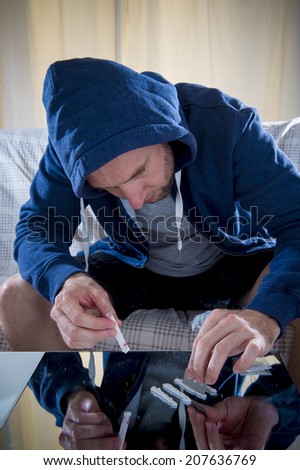 young drug addict man on hood snorting and sniffing cocaine lines on mirror with rolled banknote at home alone with moody and edgy dark studio lighting in drugs use and abuse of illegal substances