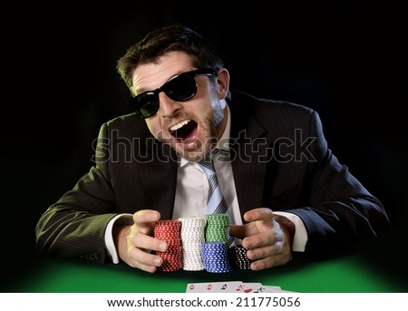 happy young attractive man grabbing poker chips after winning bet gambling on table with playing cards on green felt at Casino isolated on black background