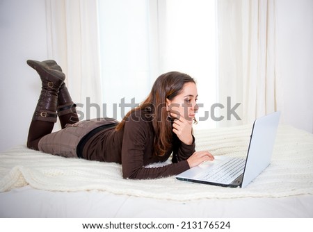 happy latin woman wearing brown jumper shirt working and using internet on her laptop laying on bed at home smiling and relaxed